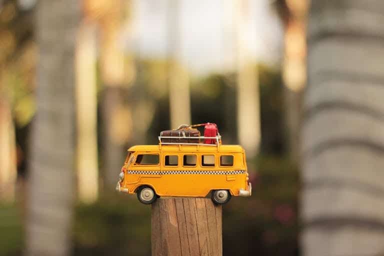 A close-up of a yellow toy campervan positioned on a wooden post in a sunny outdoor setting.
