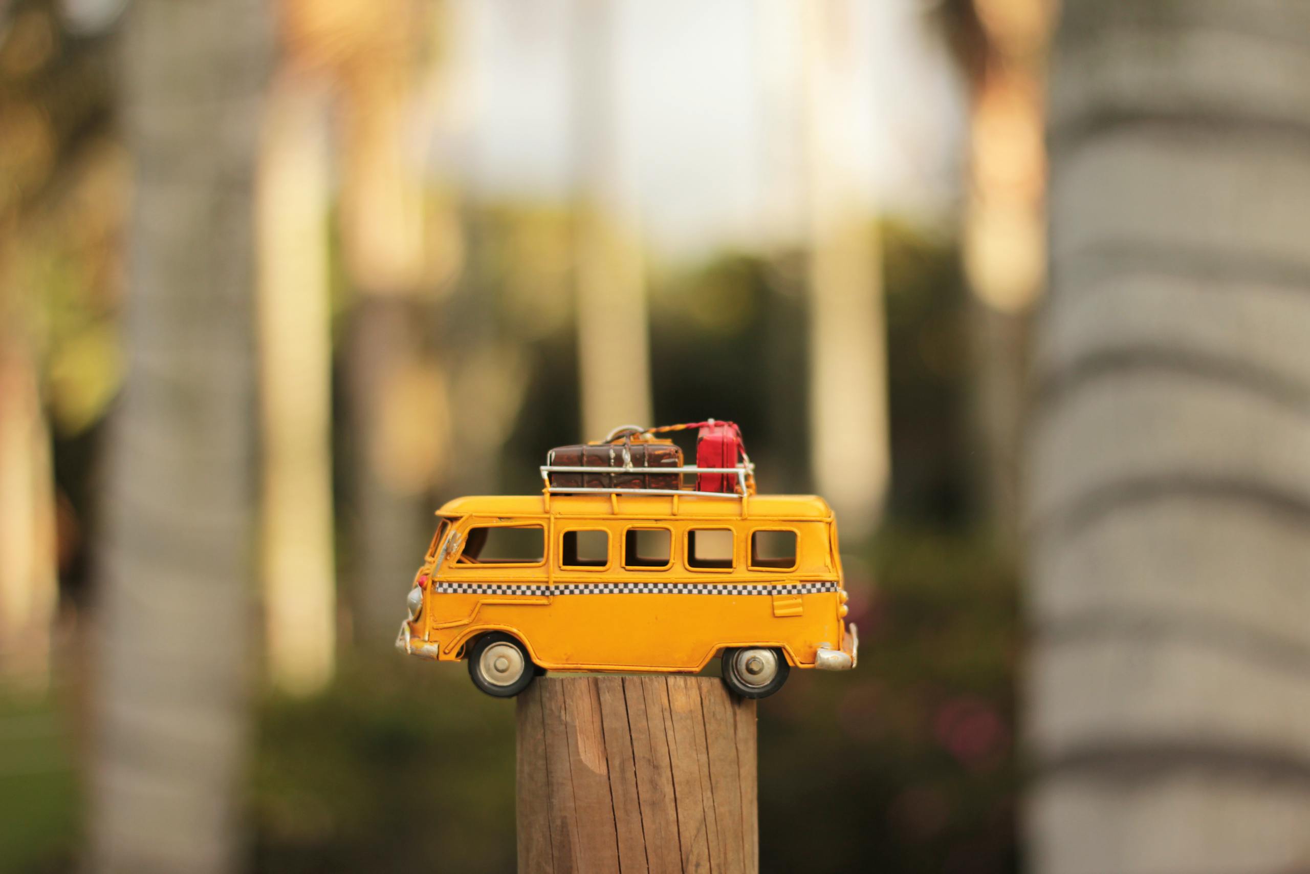 A close-up of a yellow toy campervan positioned on a wooden post in a sunny outdoor setting.