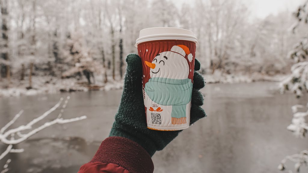 A hand in gloves holds a festive snowman coffee cup outdoors in a wintry landscape.